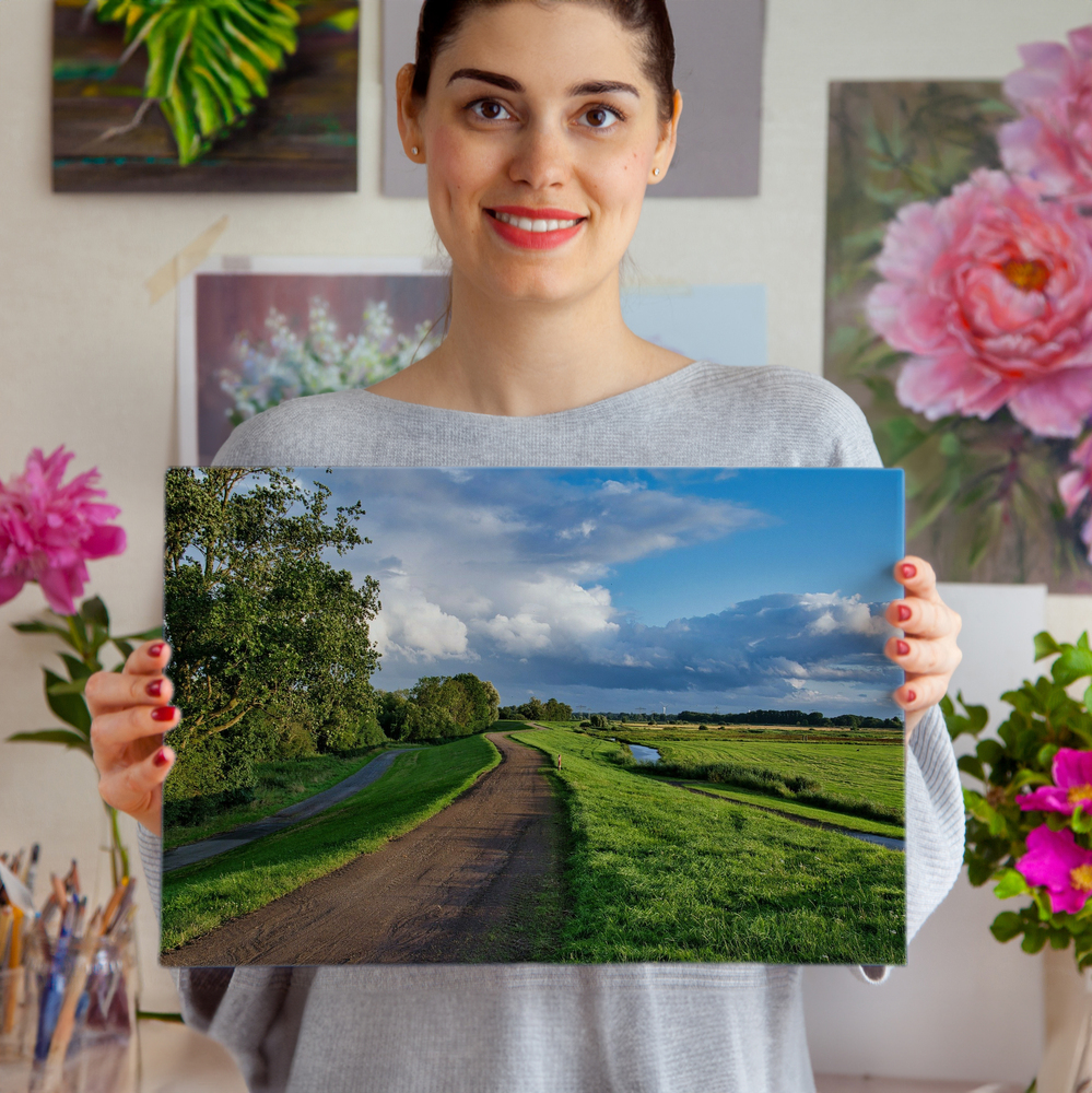 Rahmenloser Acrylglasdruck mit Feldweg, grünen Wiesen und Wolkenhimmel, vor einer Wand mit Blumenbildern