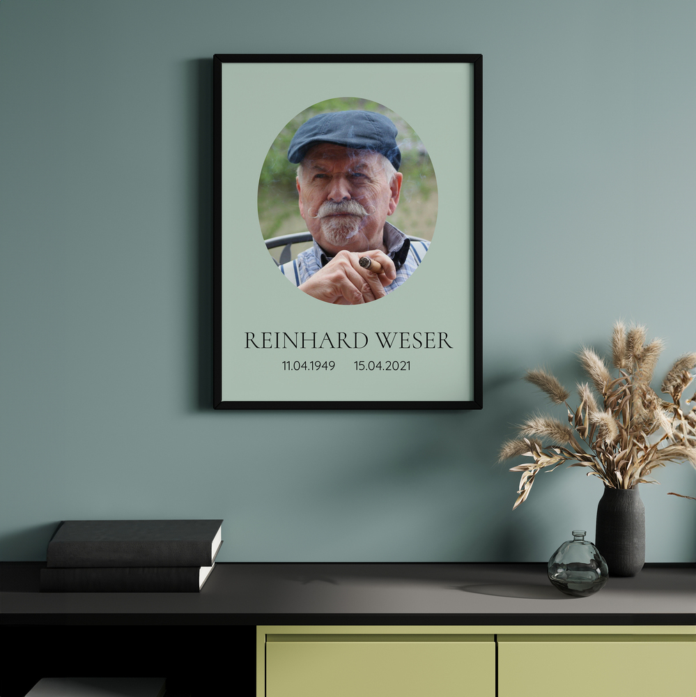Trauerfoto mit ovalem Porträt und Lebensdaten im schwarzen Rahmen über einem Sideboard neben Trockenblumen